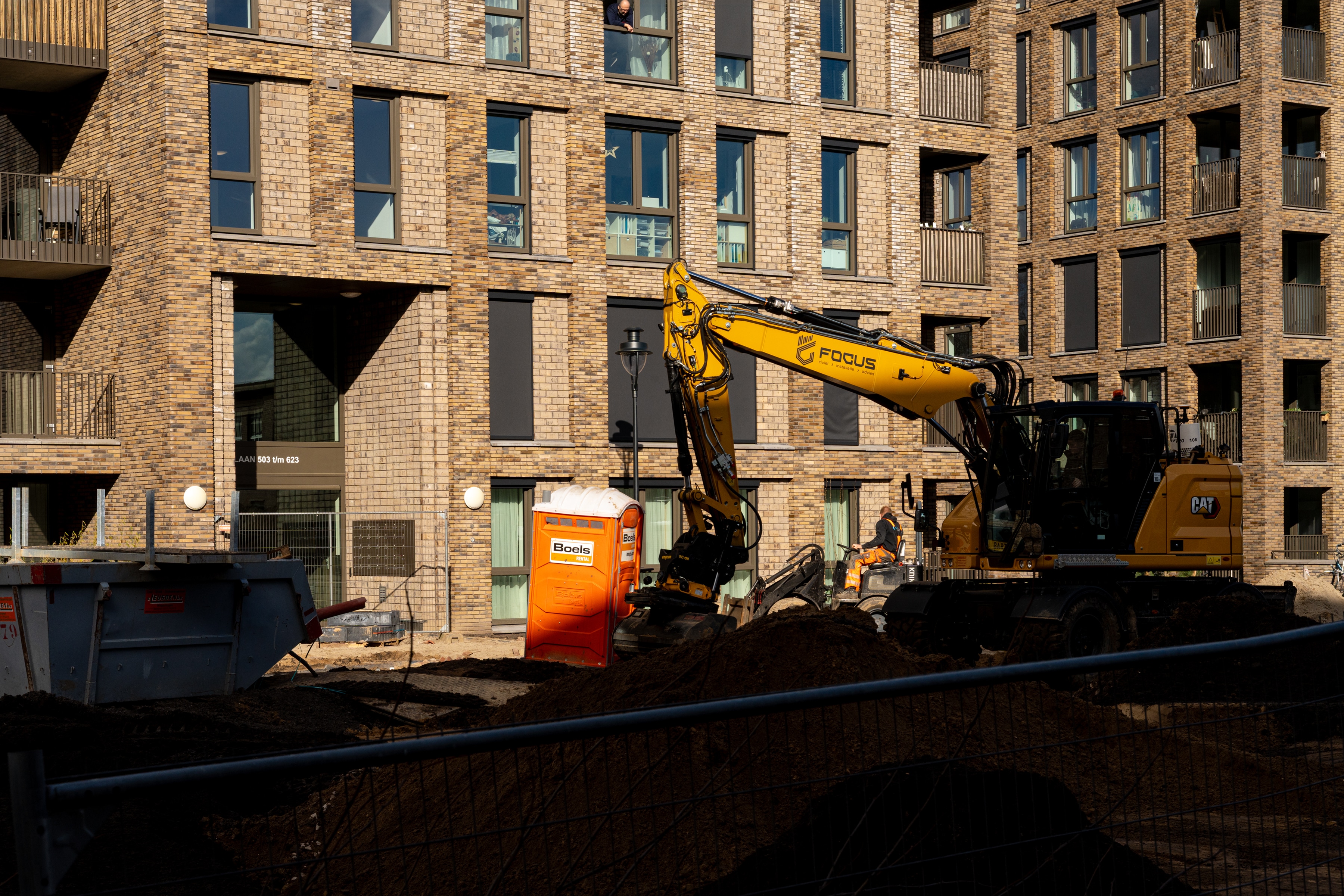 Een gele graafmachine werkt op een bouwterrein voor een modern bakstenen flatgebouw. Een oranje draagbaar toilet en hopen vuil zijn zichtbaar, met arbeiders en apparatuur op de bouwplaats.