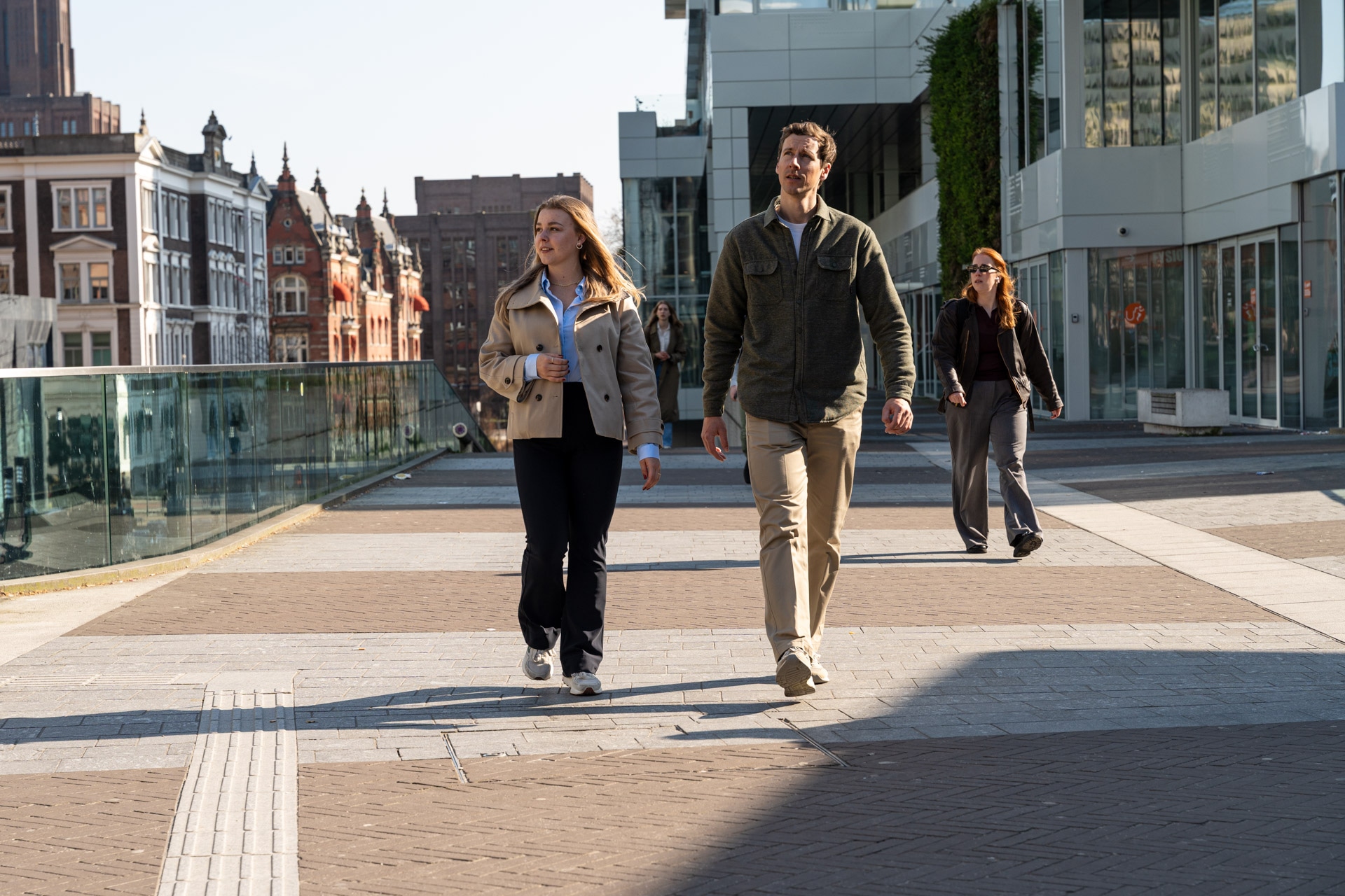 Twee mensen lopen naast elkaar op een moderne straat in de stad, terwijl een andere persoon hen volgt. De straat heeft glazen balustrades, hoge gebouwen en oudere bakstenen architectuur op de achtergrond.