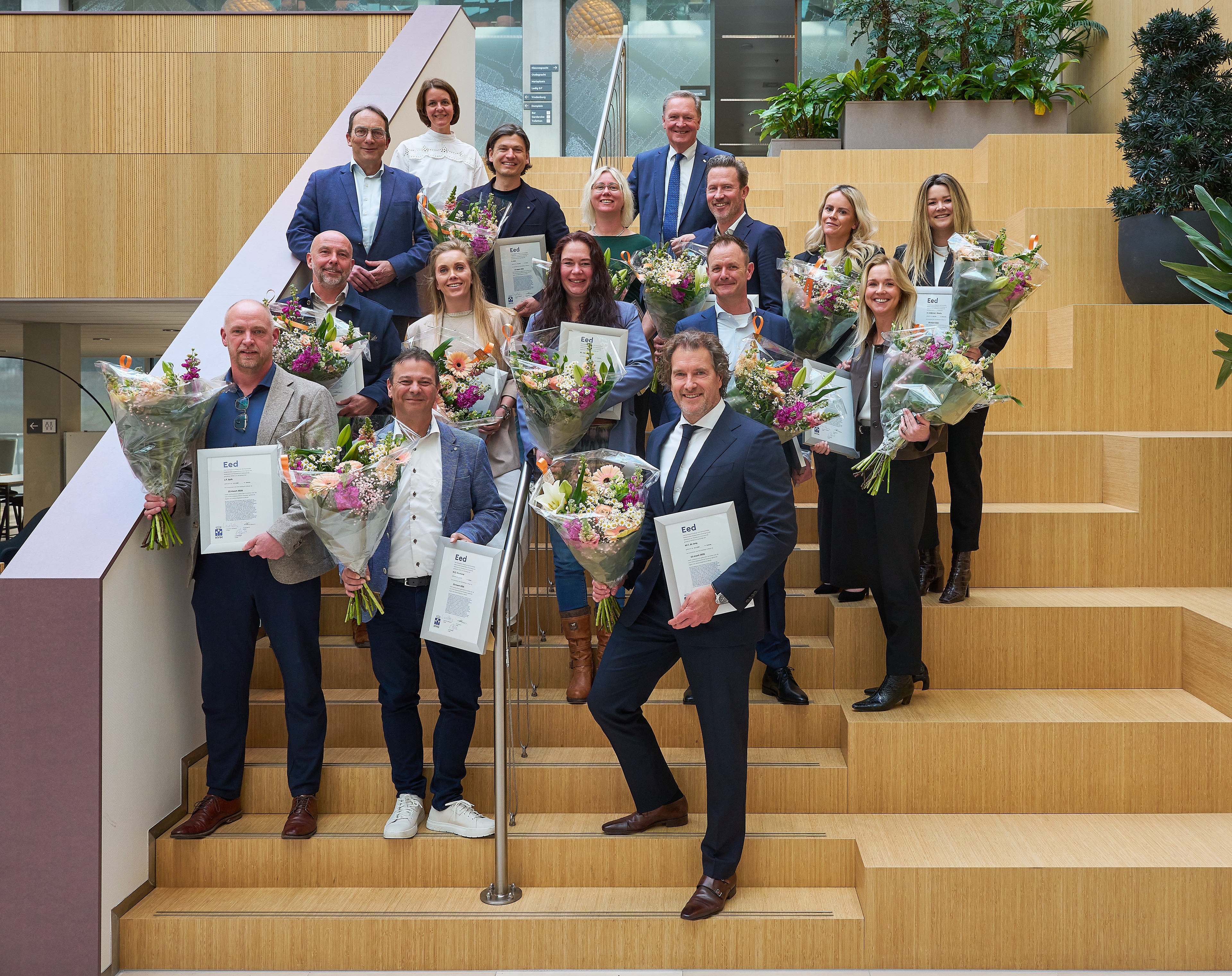 Een groep mensen staat binnen op een houten trap, elk met een boeket bloemen en een certificaat in de hand, lachend naar de camera. Er staan planten in de buurt en een paar mensen staan bovenaan de trap.