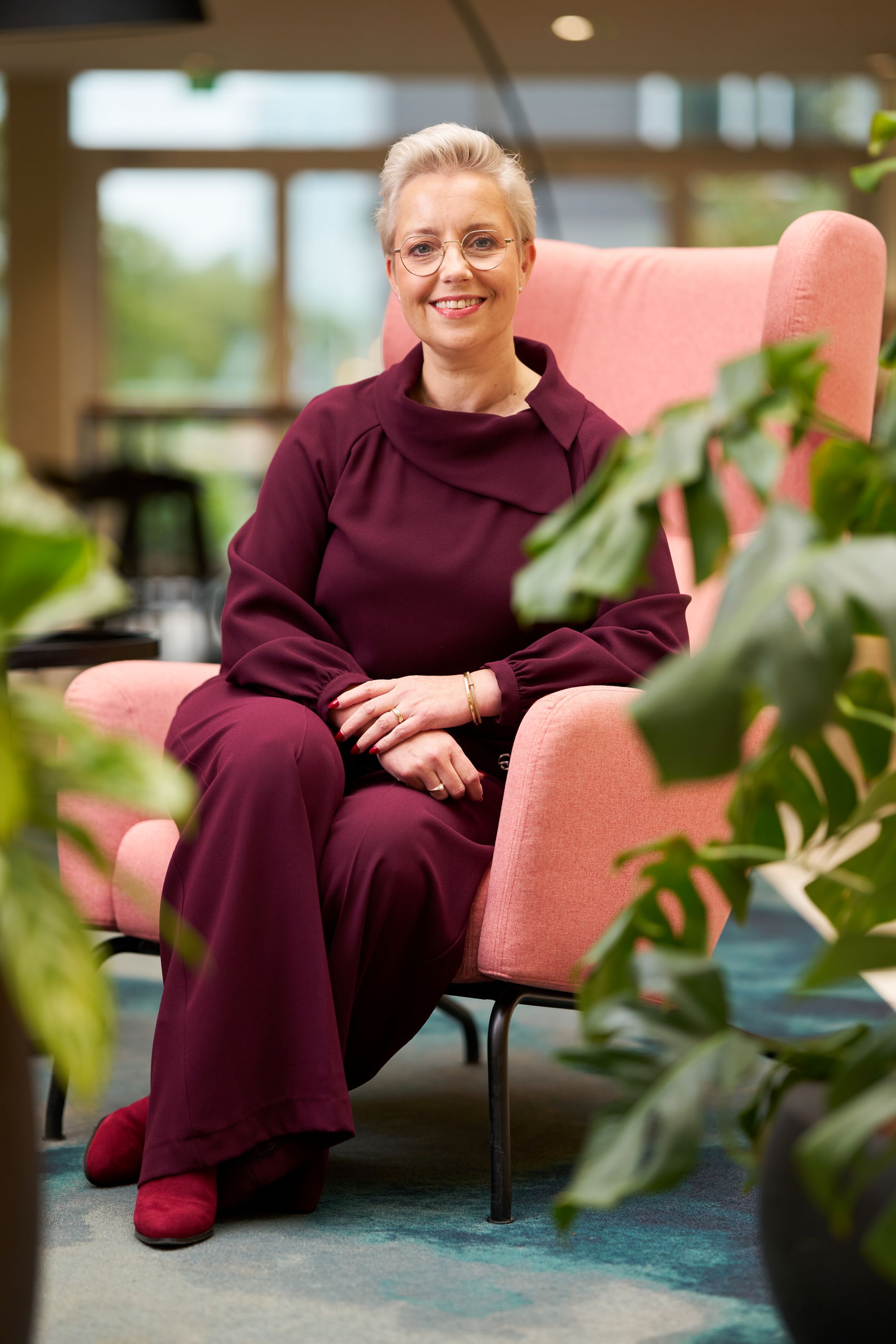Een glimlachend persoon met kort blond haar en een bril zit op een roze leunstoel, draagt een bordeauxrode outfit en rode schoenen, omringd door groene planten in een goed verlichte binnenomgeving.
